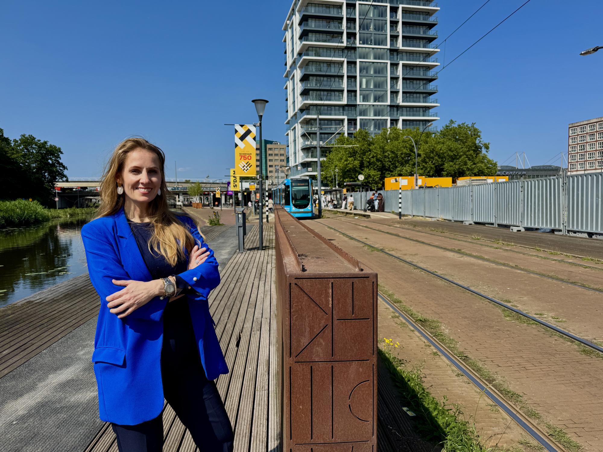 NPNO-directeur Fraukje Douwma staat met gekruiste armen glimlachend langs de trambaan, met nieuwbouw en het stationsgebied van Schiedam Oost op de achtergrond.