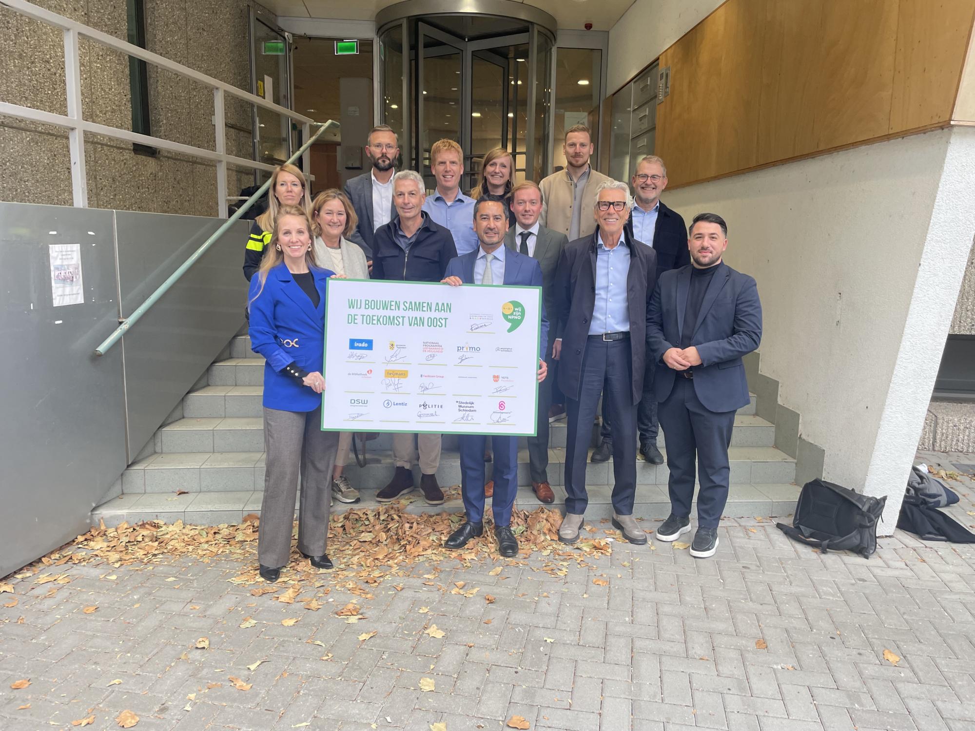 Een groep vertegenwoordigers van het Nationaal Programma staat bij de ingang van een gebouw en houdt gezamenlijk een groot bord vast waarop de samenwerkende partners van het Toekomstperspectief Oost staan vermeld.